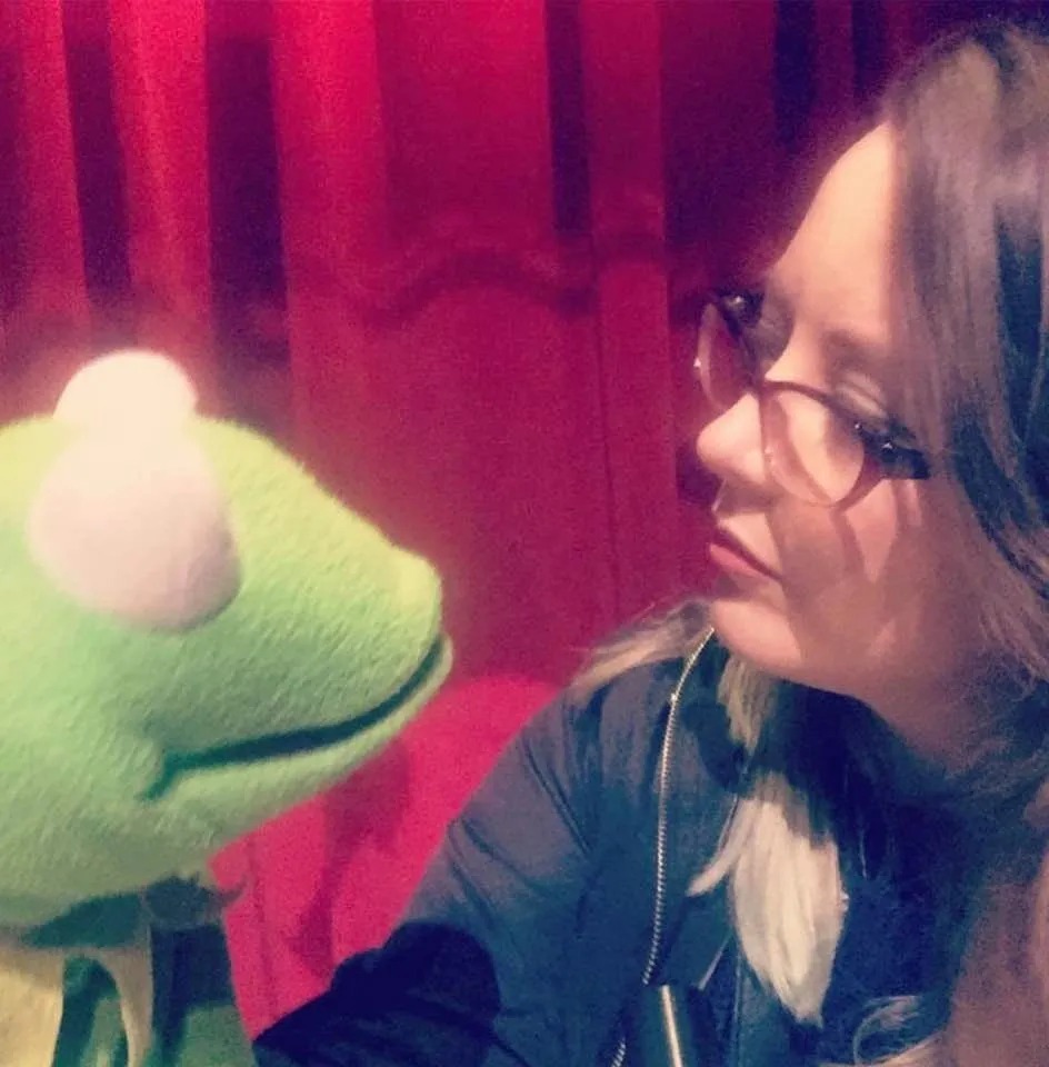 Hana is wearing glasses and looking at a Kermit the Frog puppet. The background is a red curtain, and the two are facing each other.