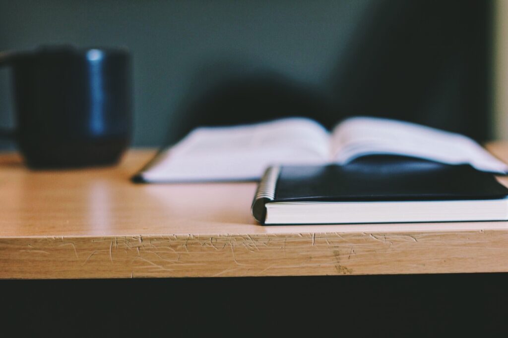 A notebook and a mug sitting on a wooden table. The background is blurred, with an open book out of focus.