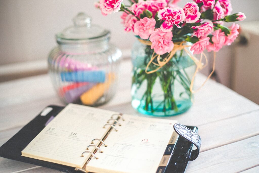 An open planner on a wooden table with a glass jar filled with coloured chalk and a vase of pink carnations in the background.