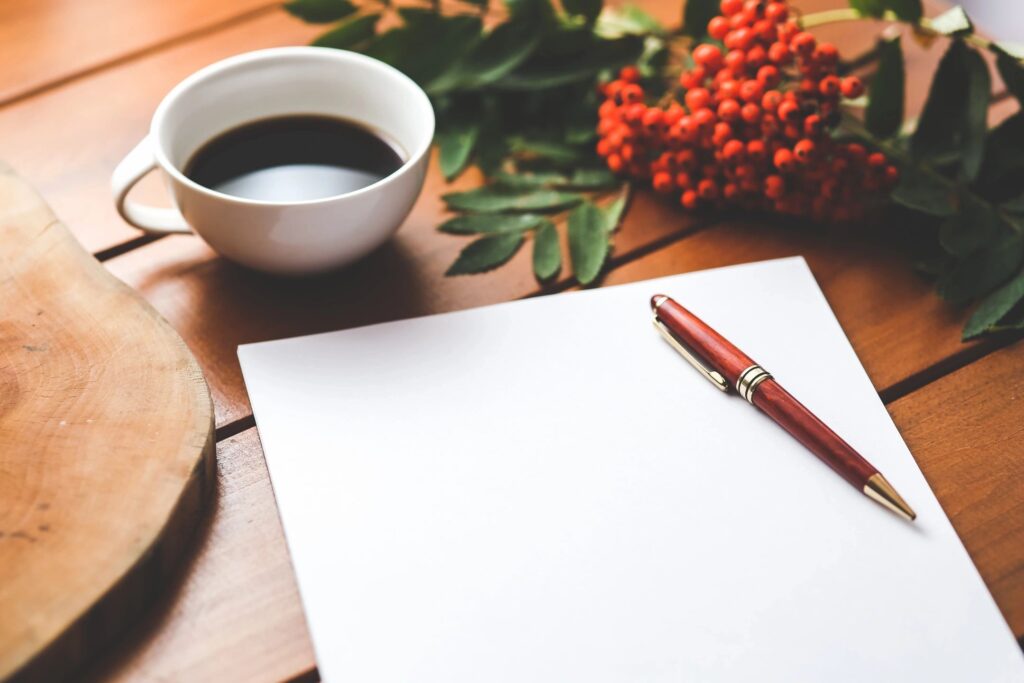 A cup of black coffee, a blank sheet of paper, and a red ballpoint pen rest on a wooden table, with red berries and green leaves in the background.