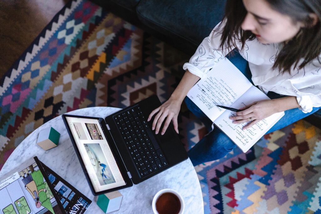 A woman working on a laptop while taking notes in a notebook. She is sitting in front of a table with a colourful patterned rug underneath and a cup of tea beside her.