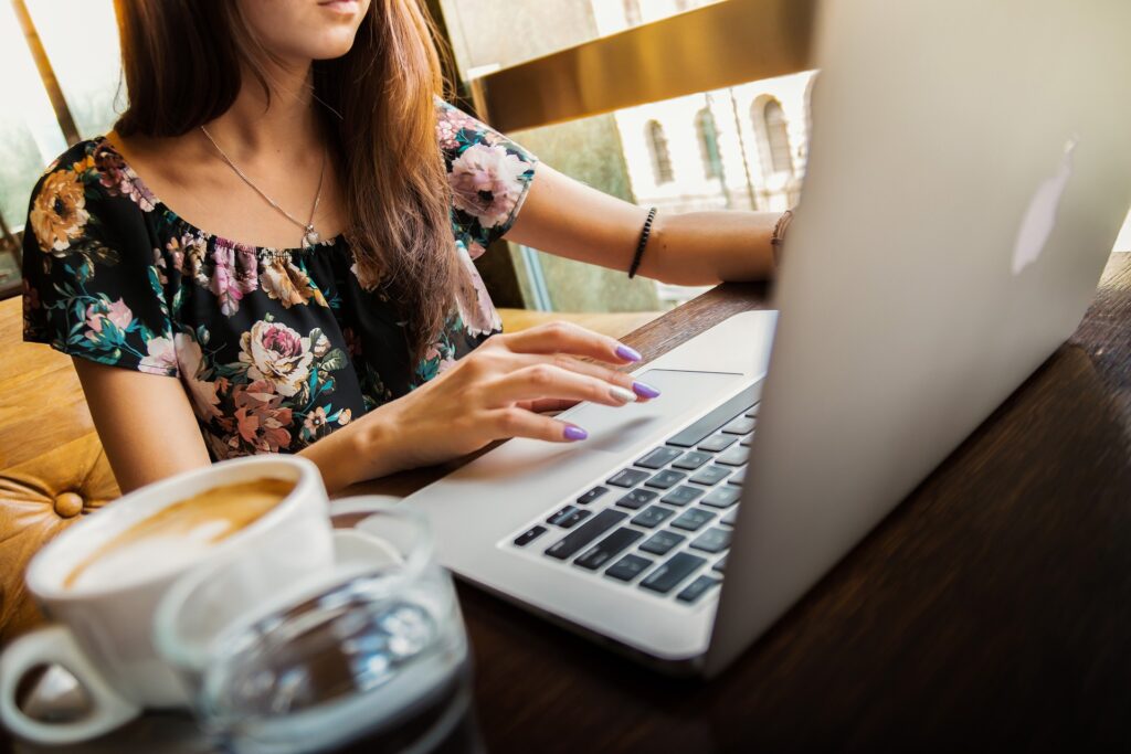 A woman typing on a laptop, with a cup of coffee and water on the table. She is wearing a floral-patterned shirt, and the background shows windows with sunlight.