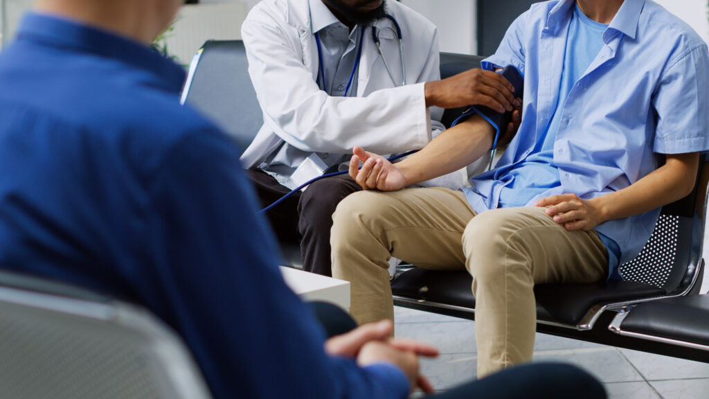 A doctor in a white coat checks a patient's blood pressure using a cuff, while another person sits nearby. The patient is sitting in a chair wearing a light blue shirt.