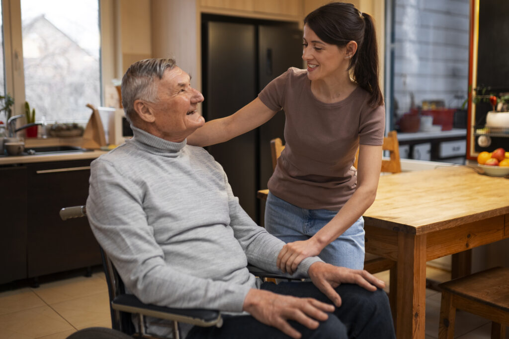 A smiling elderly man in a wheelchair engages with a young woman standing beside him in a cosy kitchen setting. The woman has her hand on his shoulder, showing care and connection.