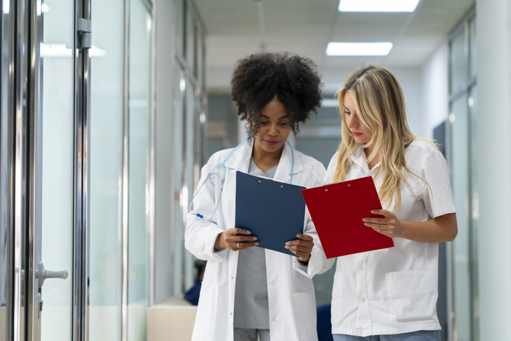 Two female doctors, one with curly hair and the other with straight blonde hair, stand in a hallway, discussing medical charts they are holding.