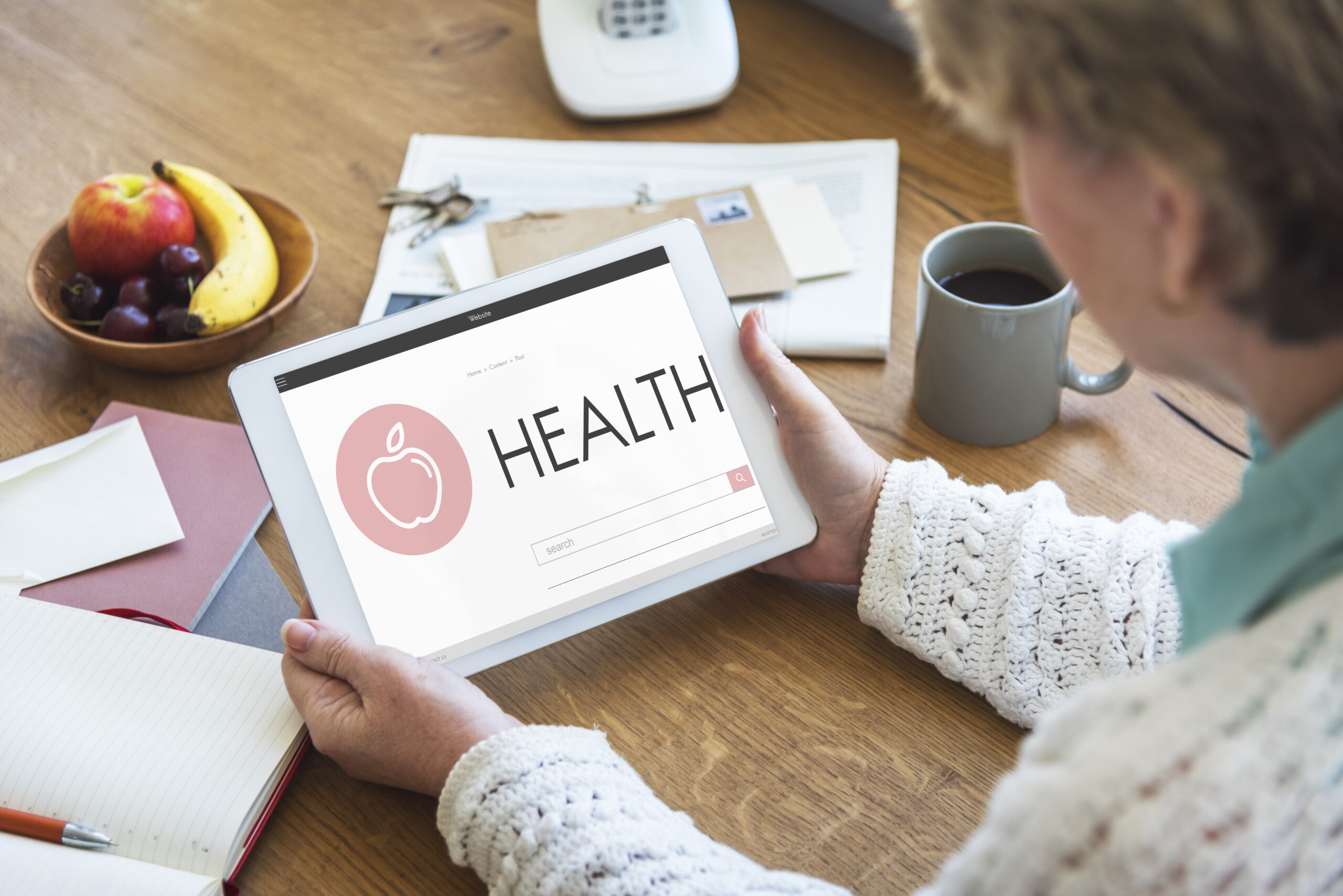 A person holds a digital tablet displaying the word "Health" with an icon of an apple on the screen. The table is cluttered with papers, a bowl of fruit, and a cup of coffee.