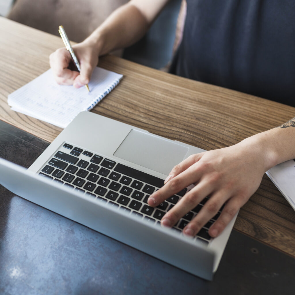 A person writes notes in a notepad while using a laptop on a wooden table, concentrating on their work.