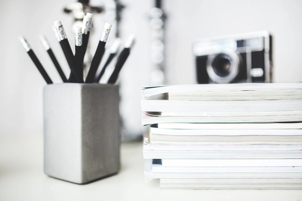 A cup filled with black pencils, with a stack of books and a blurred background that includes a camera.