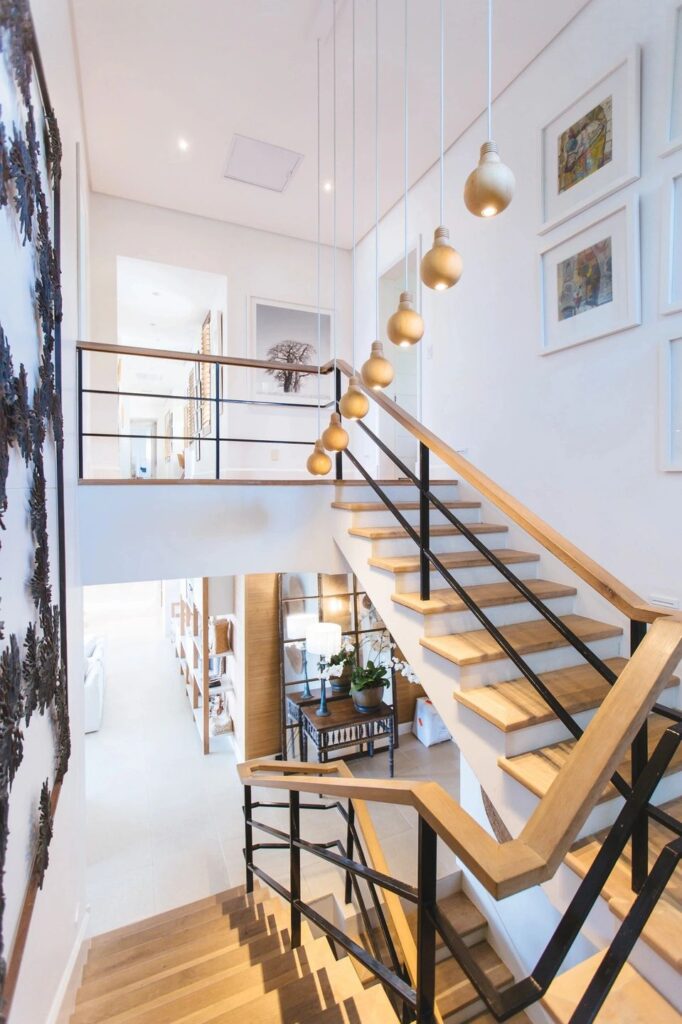 A contemporary staircase with wooden steps and black railings. Hanging pendant lights are arranged above the staircase, and there are framed pictures on the walls.
