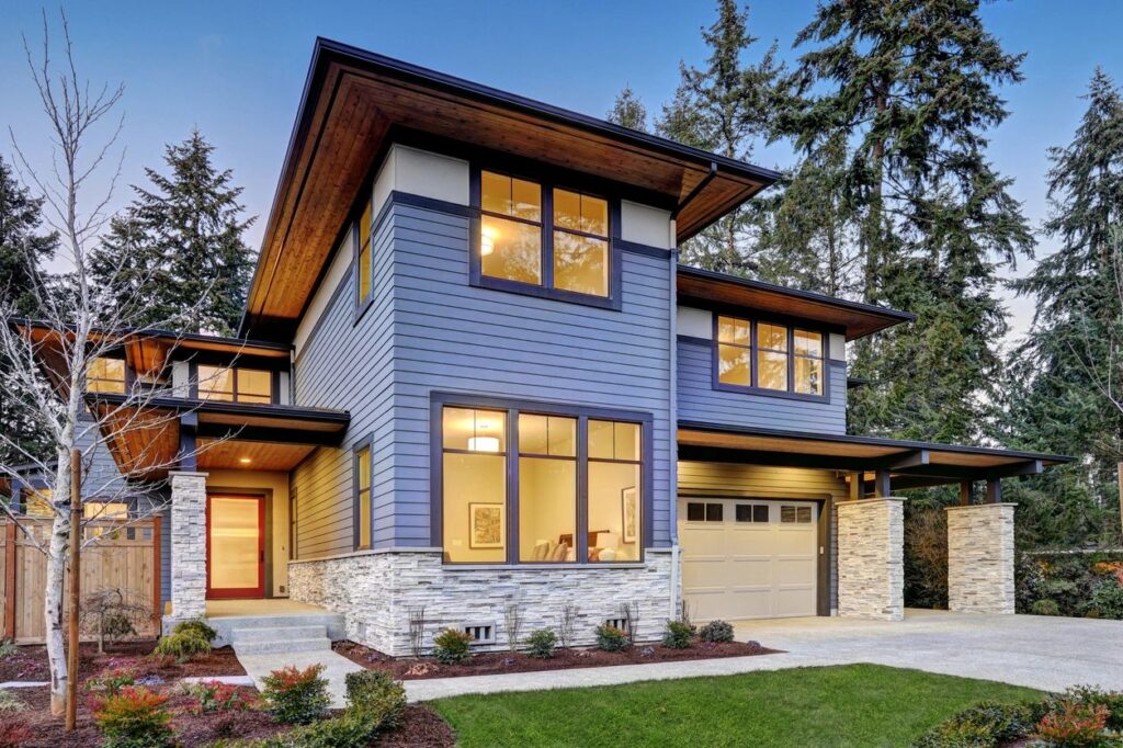 A modern two-story house with blue siding, large windows, and a garage, surrounded by trees and landscaping.