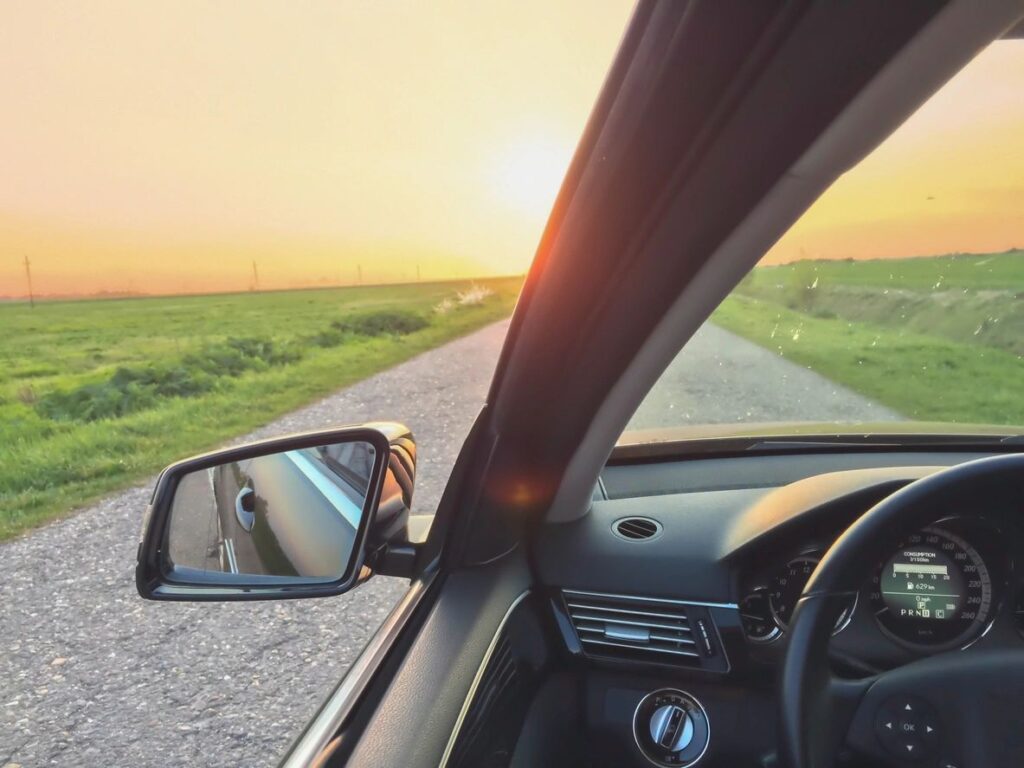 The interior view of a car driving down a rural road with green fields on both sides, during a sunset. The car's rear view mirror and dashboard are visible.