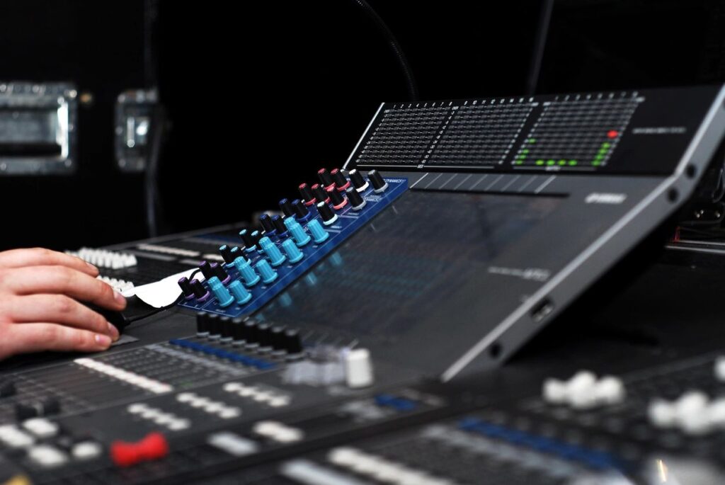 A hand adjusting coloured dials on a large soundboard or mixing console, with various buttons and sliders visible.