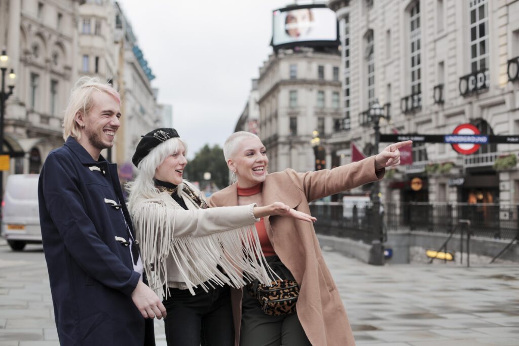 Three people in London, smiling and pointing at something off-camera. One is wearing a tan coat, another in a fringed jacket, and the third in a dark coat. They are standing near an Underground entrance.