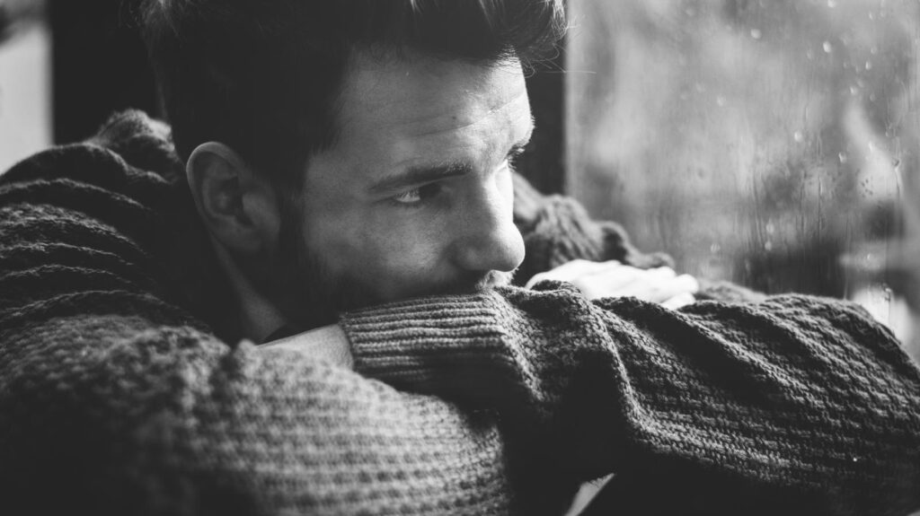 A black-and-white photo of a man wearing a sweater, resting his chin on his arms and looking out of a window on a rainy day.