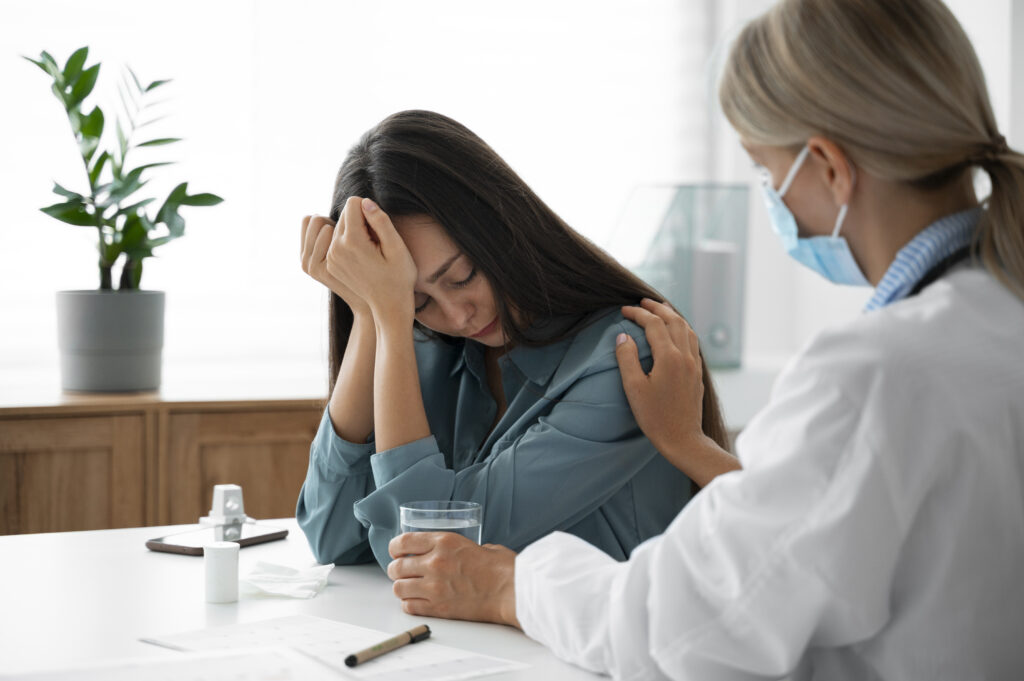 A female doctor wearing a face mask consoles a distressed woman sitting at a table holding a glass of water. A potted plant and medical documents are on the table.