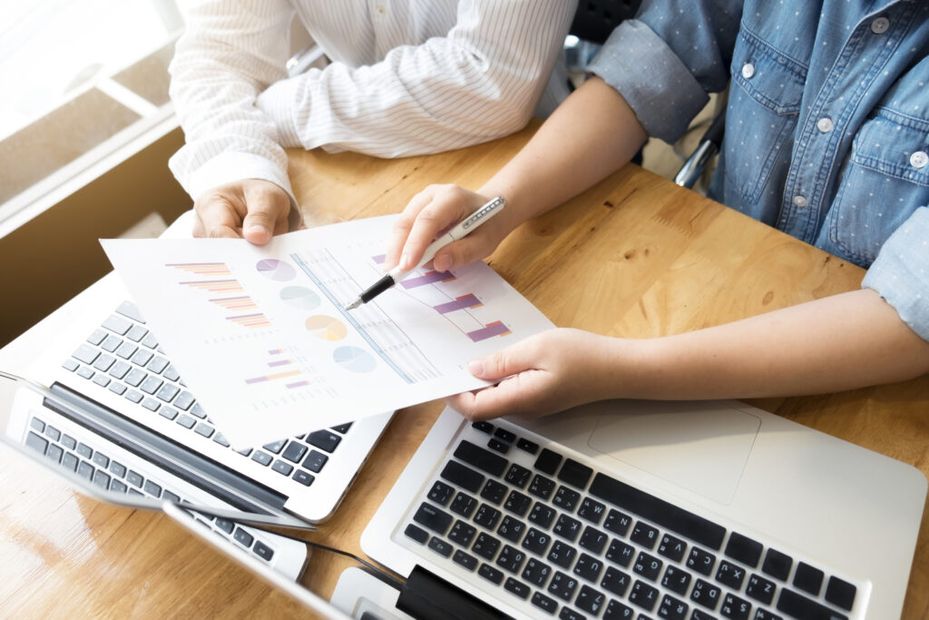 Two people looking at a document filled with colourful graphs and charts while sitting at a table with laptops in front of them.