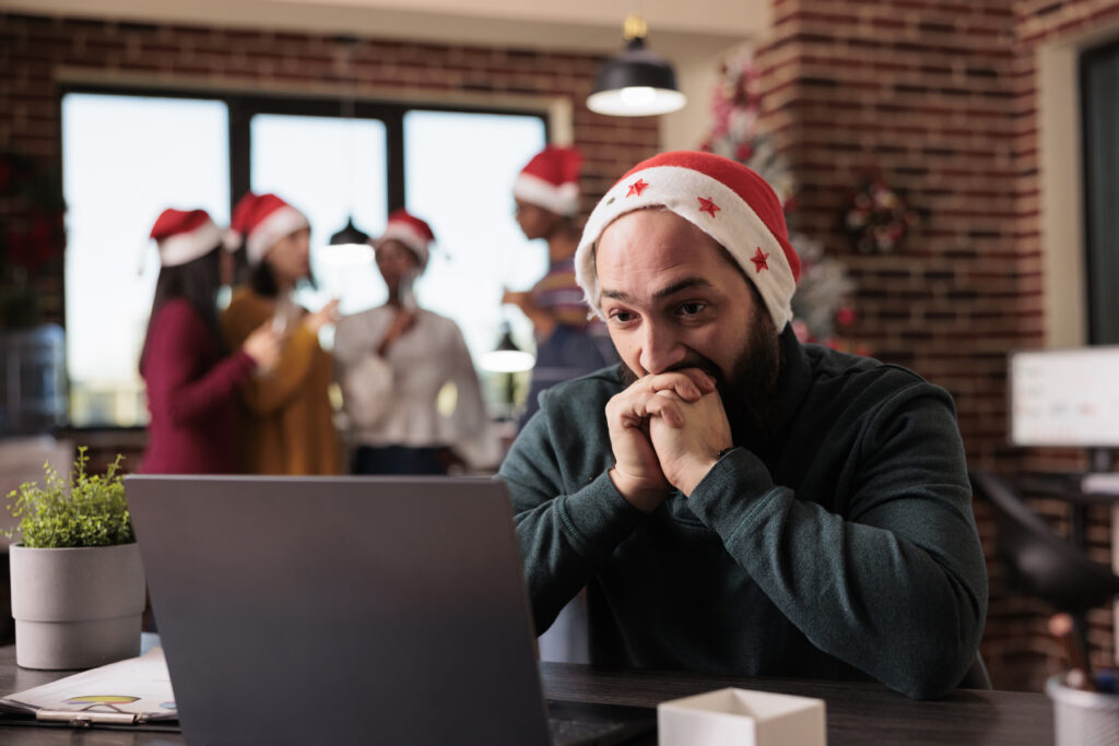 A man sitting at a desk, wearing a Christmas hat, looking at a laptop with a stressed expression. In the background, coworkers in festive hats are chatting and celebrating