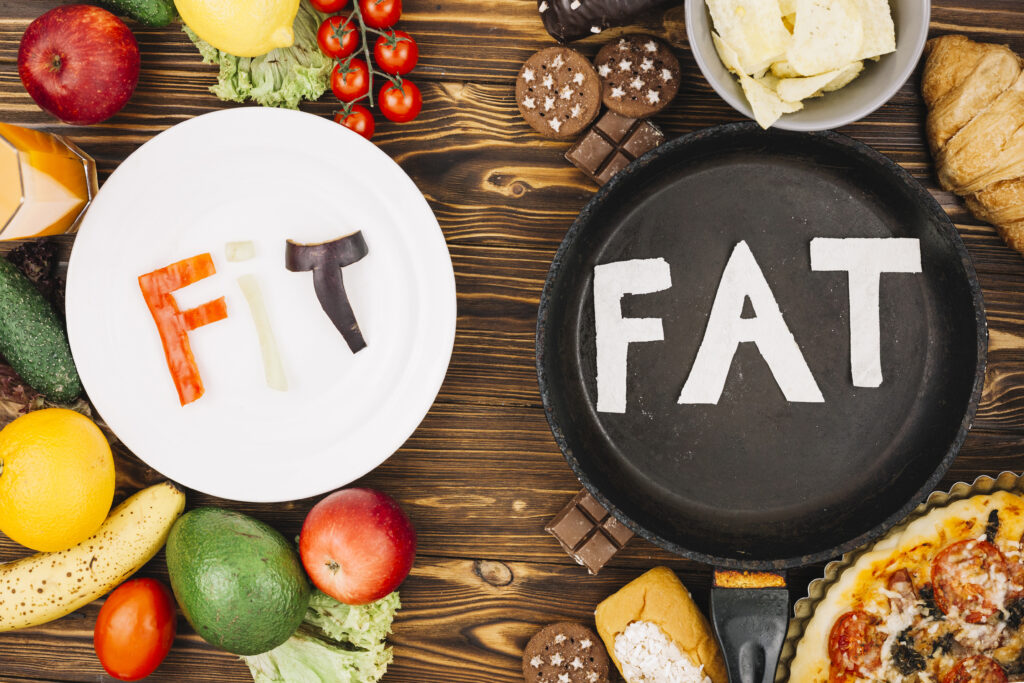 On the left is a white plate with the word "Fit" spelled out in vegetables. Around the plate are fruits and vegetables. On the right, a black pan with the word "Fat" carved out of fat, surrounded by high-calorie foods.