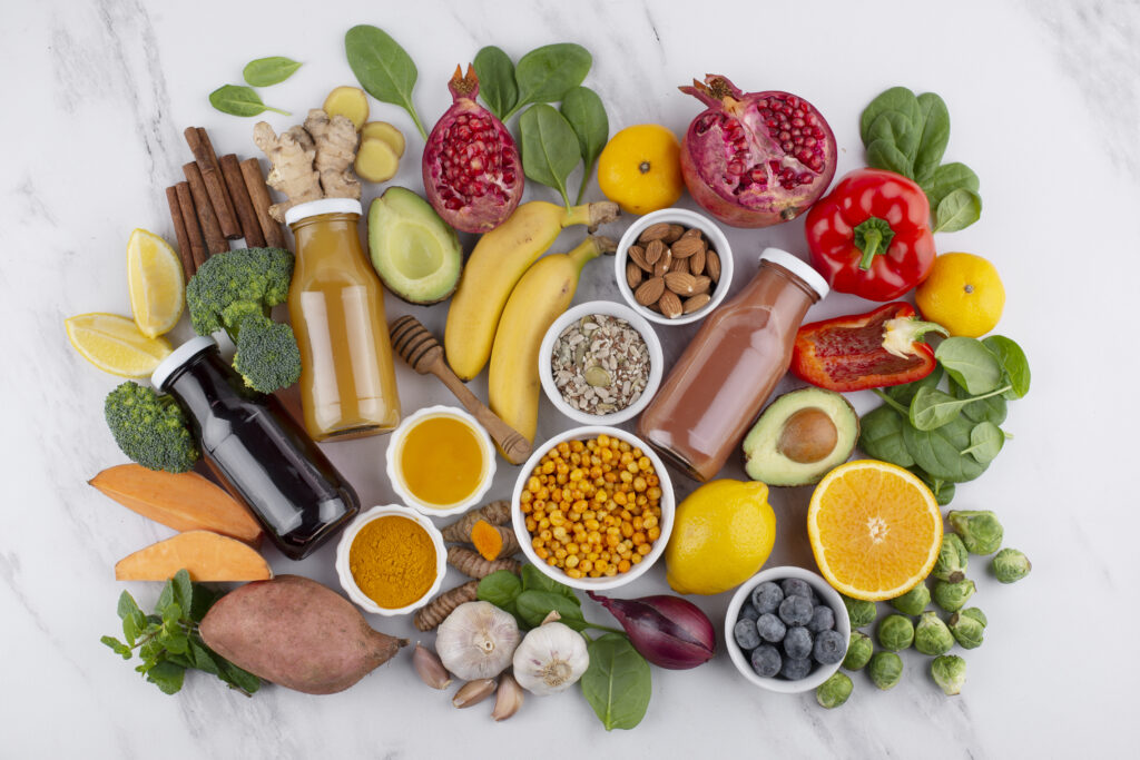 A flat lay of healthy foods and drinks, including fruits, vegetables, nuts, and juices. Items like pomegranates, bananas, broccoli, sweet potatoes, and honey are arranged on a white background