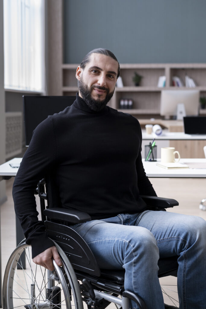 A man in a black turtleneck and jeans sits in a wheelchair, smiling at the camera. He is in a modern office setting with a desk and shelves in the background.