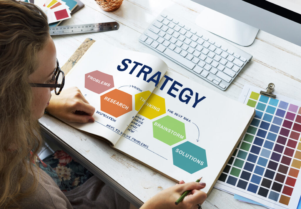 A person sitting at a desk looking at an open book labeled "Strategy" with sections for "Problems," "Research," "Thinking," "Brainstorm," and "Solutions." A color palette and keyboard are visible in the background.