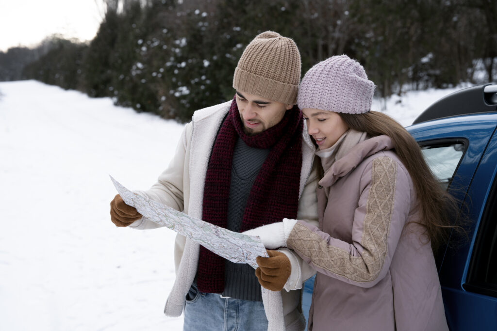 A couple dressed in winter clothing is standing outdoors in a snowy landscape, looking at a map together. They are both wearing knit hats and cozy jackets, with a blue car beside them.