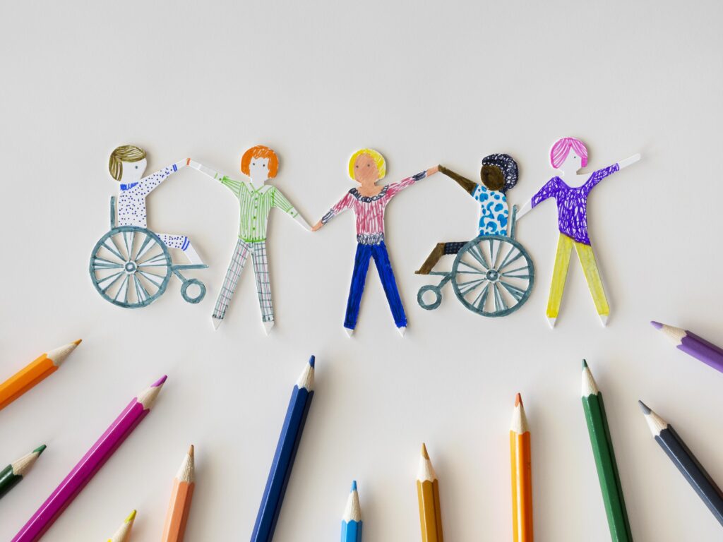 A colorful drawing of five diverse figures holding hands, including two figures in wheelchairs. The drawing is surrounded by colored pencils.