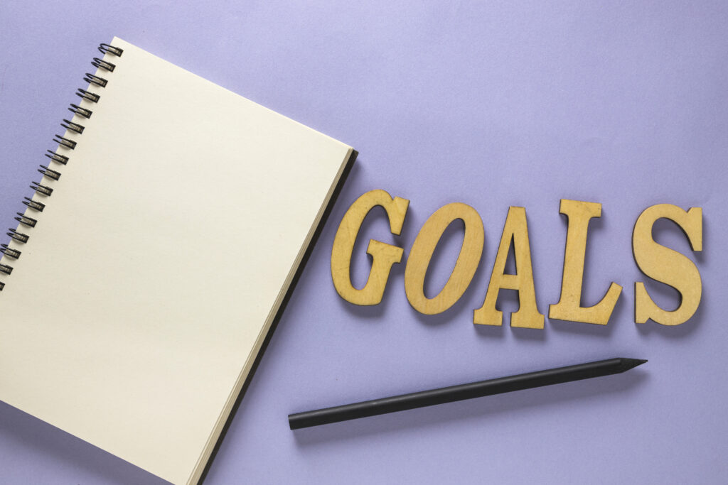 A blank notebook with wooden letters spelling "GOALS" arranged next to it, placed on a light purple background