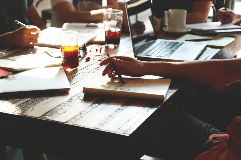 A group of people working at a table with laptops, notebooks, and drinks. One person is writing in a notebook, and another is using a laptop. Glasses of iced beverages are visible on the table.
