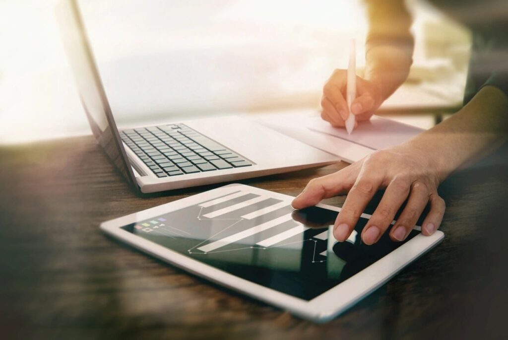 A person using a tablet with a graph displayed on the screen, while also writing on a notepad. A laptop is open on the desk beside them.