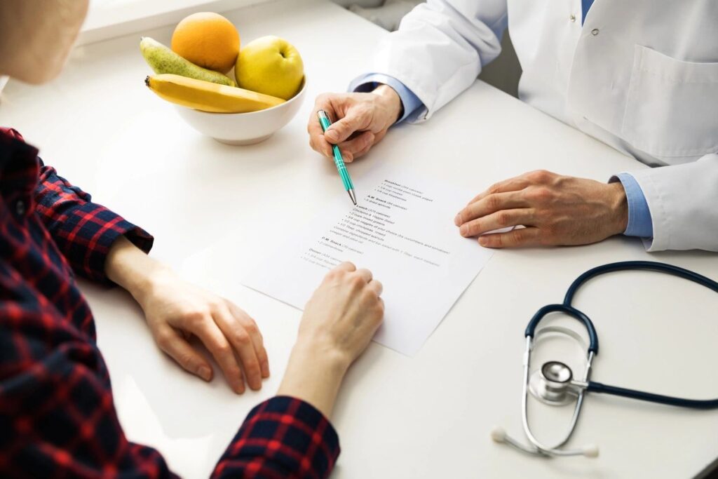 A doctor and a patient discussing a medical document on a white table. A bowl of fruit and a stethoscope are visible nearby.