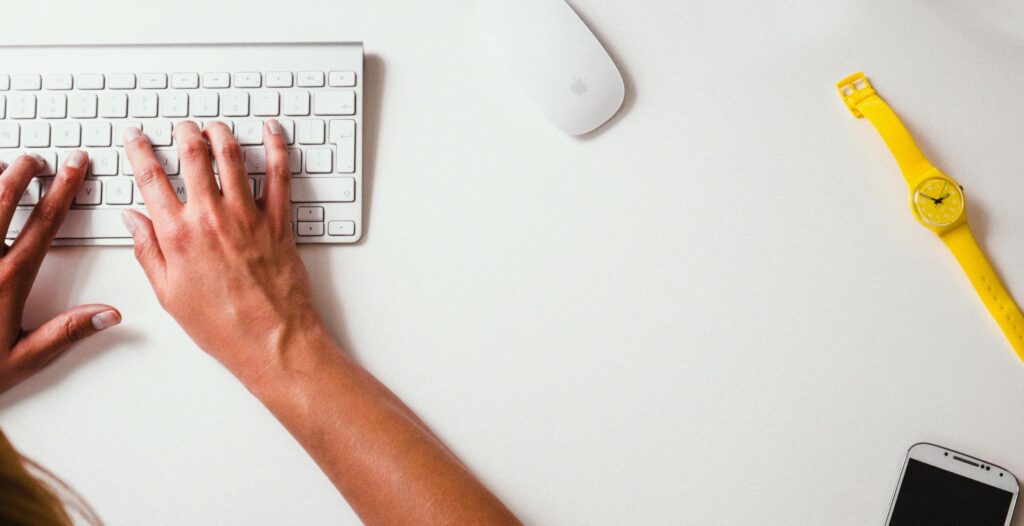 A top-down view of a person's hands typing on a white keyboard. A white wireless mouse is next to the keyboard, and a yellow wristwatch and smartphone are placed nearby.