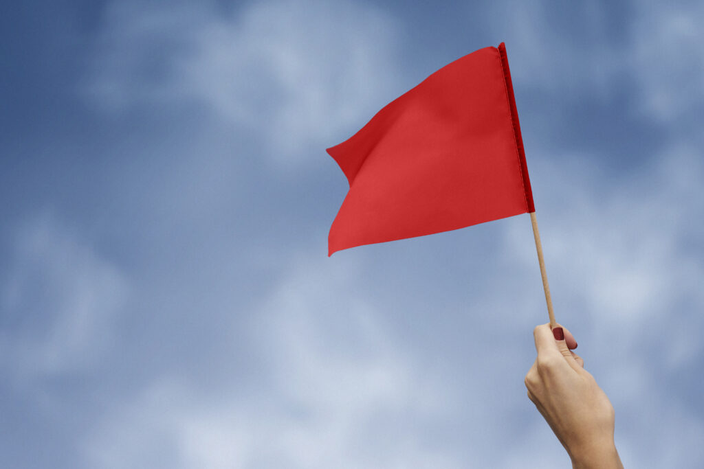 A red flag being held up by a hand, against a backdrop of a blue sky with clouds.