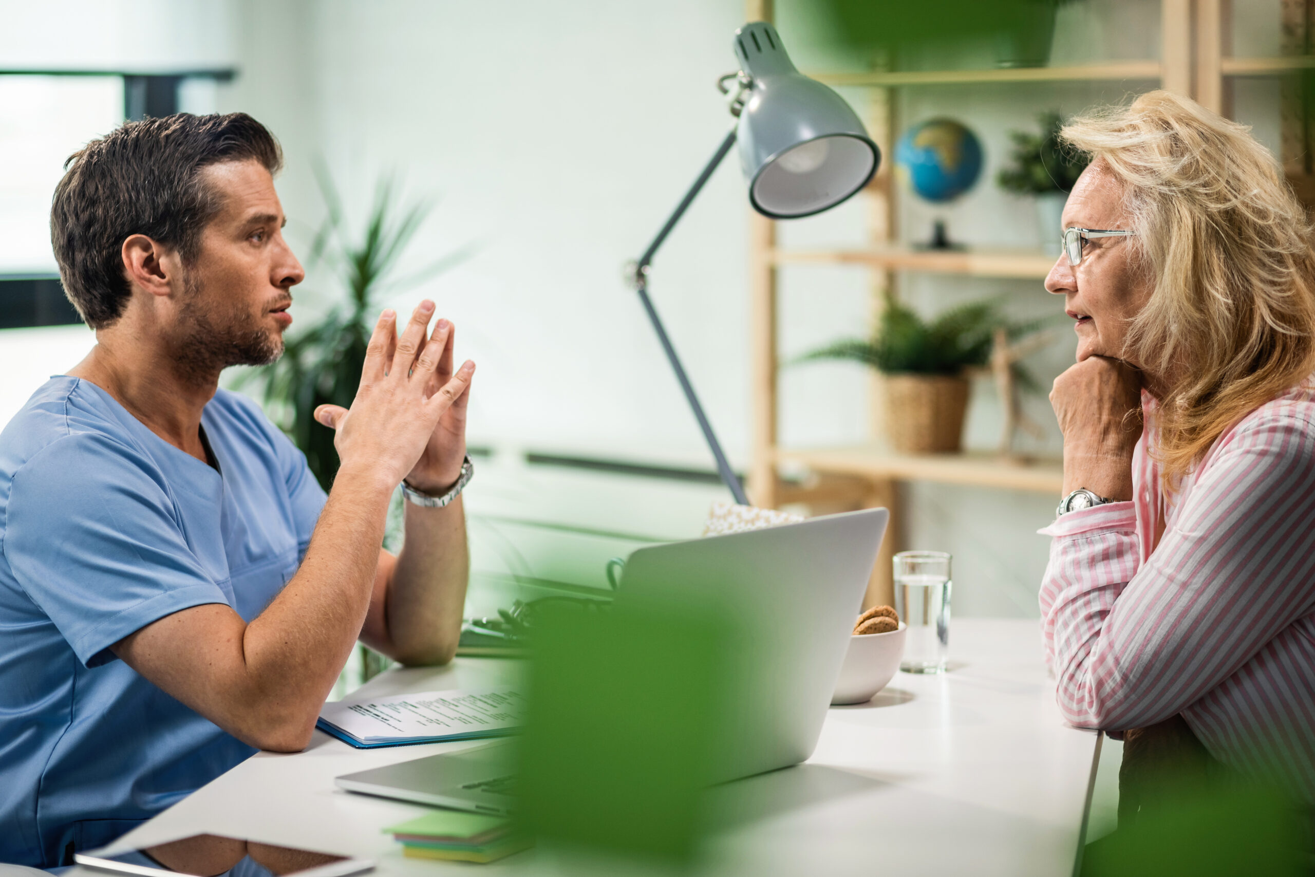 A male healthcare professional in scrubs speaks with an older woman in a striped shirt during a consultation in a bright, well-decorated office.