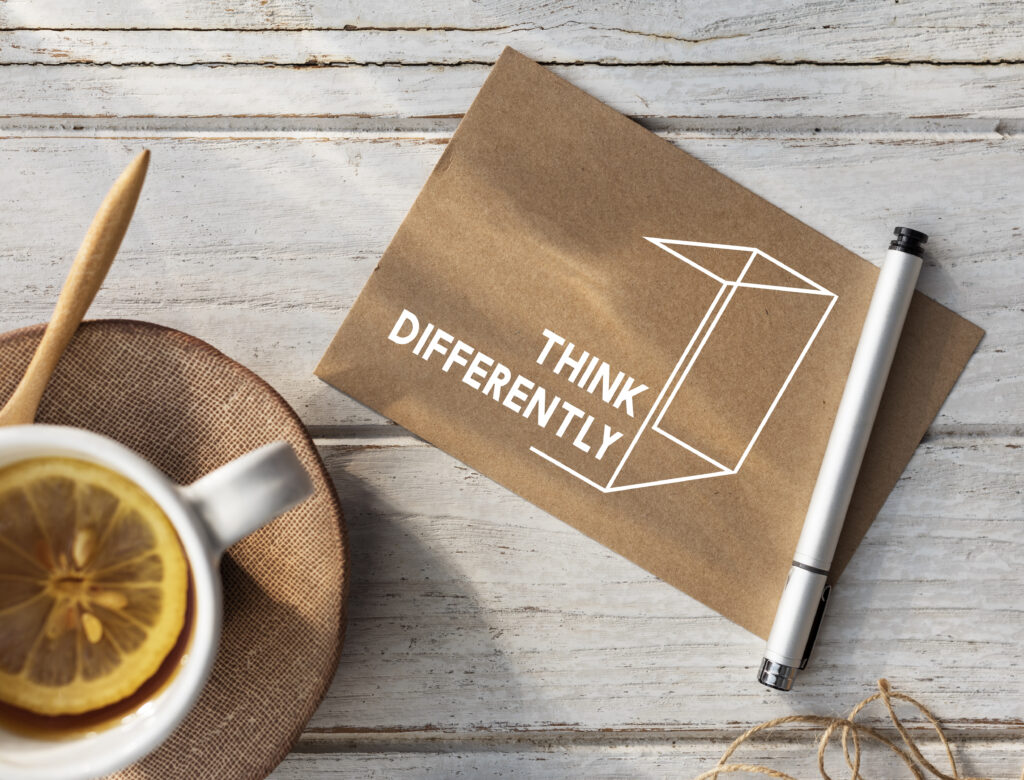 A cup of tea with a lemon slice next to a brown envelope with the words "Think Differently" printed in white. A pen rests on the envelope, and everything is set on a rustic white wooden table.