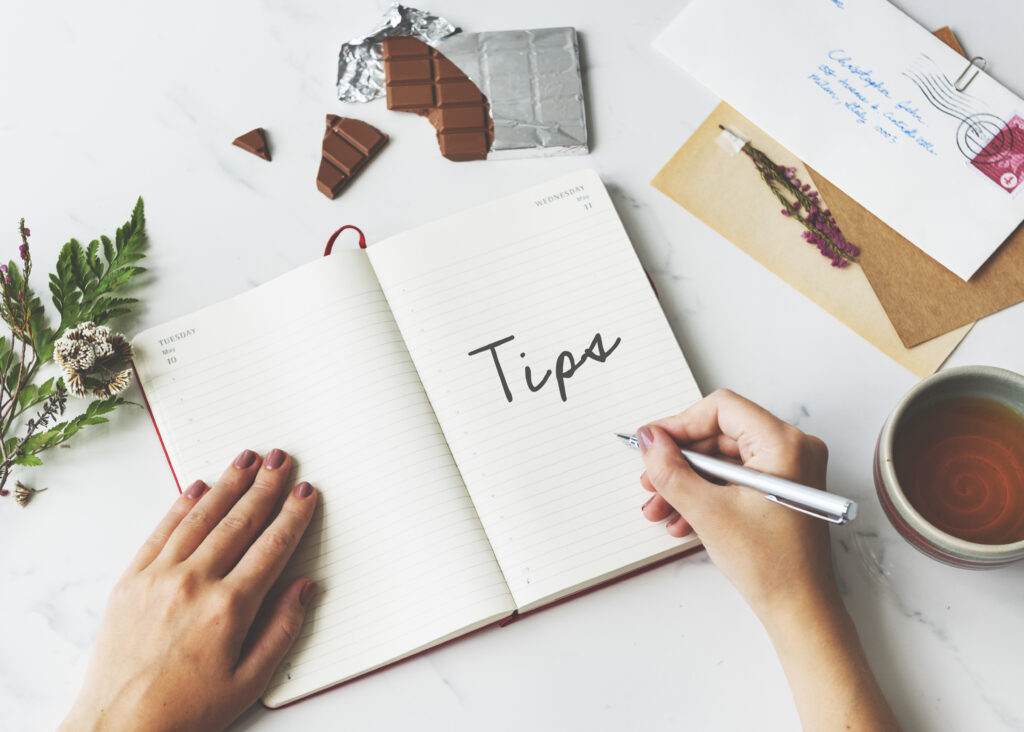 A notebook open on a table with the word "Tips" written inside. A person is writing, with chocolate, tea, and flowers arranged on the table