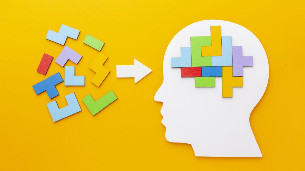 A cutout silhouette of a human head with colorful puzzle pieces inside the brain area, symbolizing thinking or problem-solving. Loose puzzle pieces and an arrow point toward the head, set against a bright yellow background.