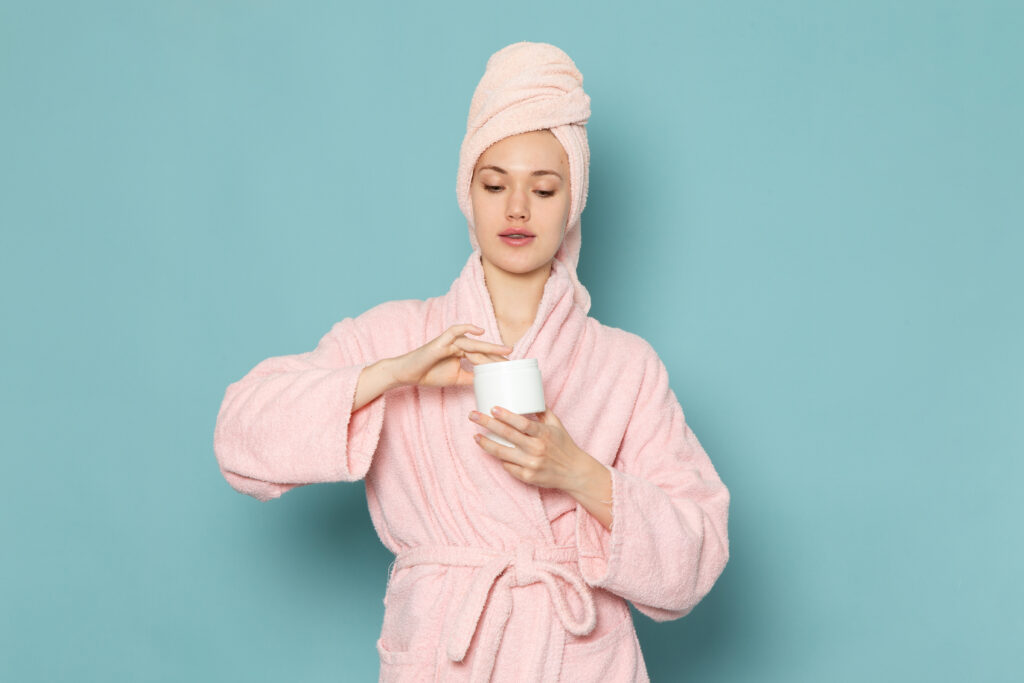 A woman in a pink bathrobe and towel holding a jar of cream against a blue background, as if applying it