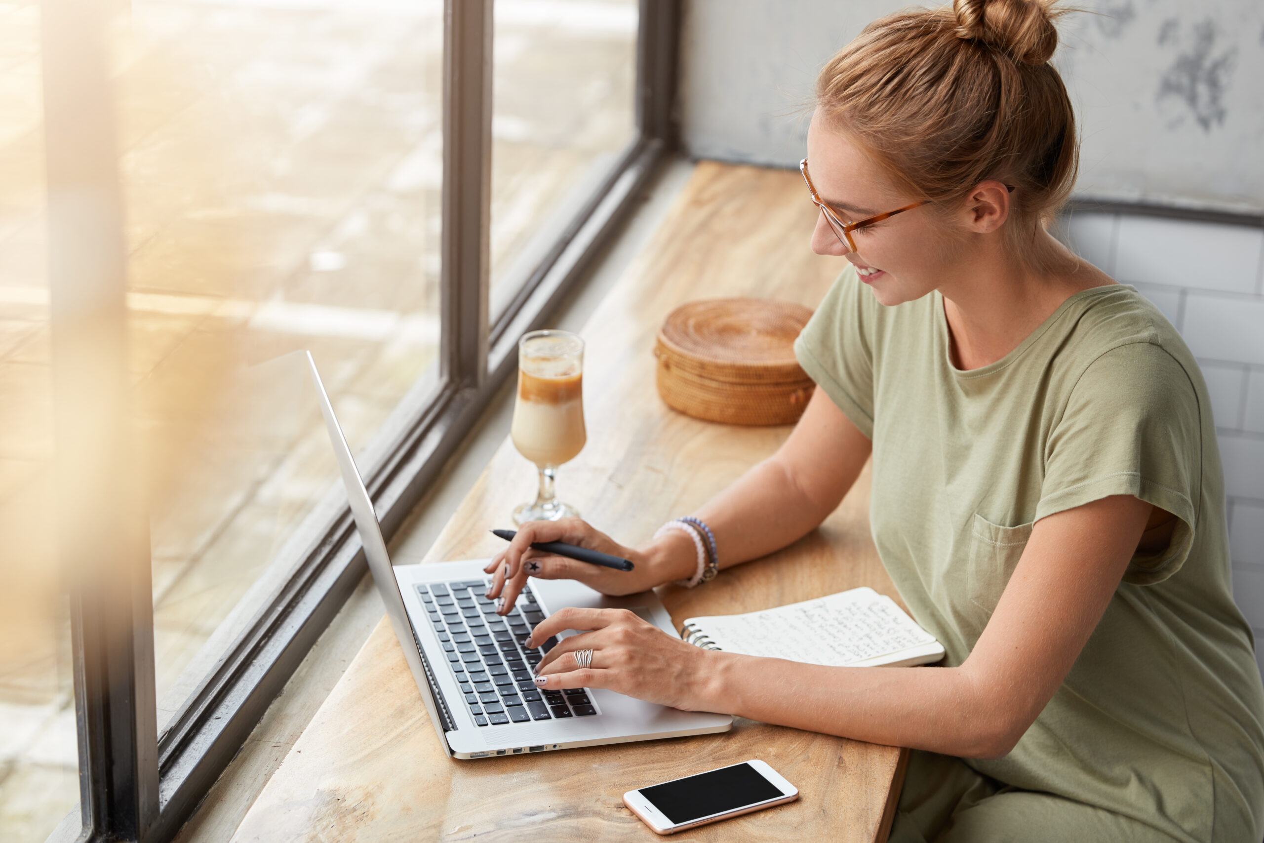 A young woman sitting at a café table, working on a laptop with a notepad beside her. She has a cup of iced coffee and is wearing glasses, smiling as she types.