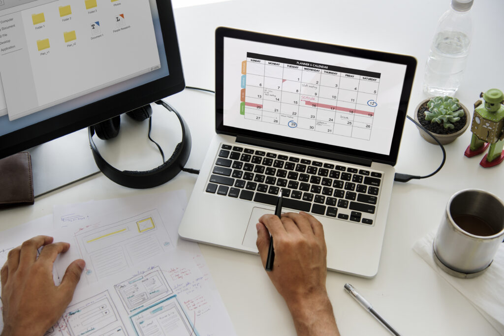 A person working on a laptop with a calendar open on the screen, surrounded by paperwork and a second monitor.