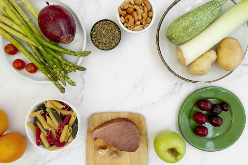 A flat lay of various fresh foods like vegetables, nuts, and pasta on a white surface.