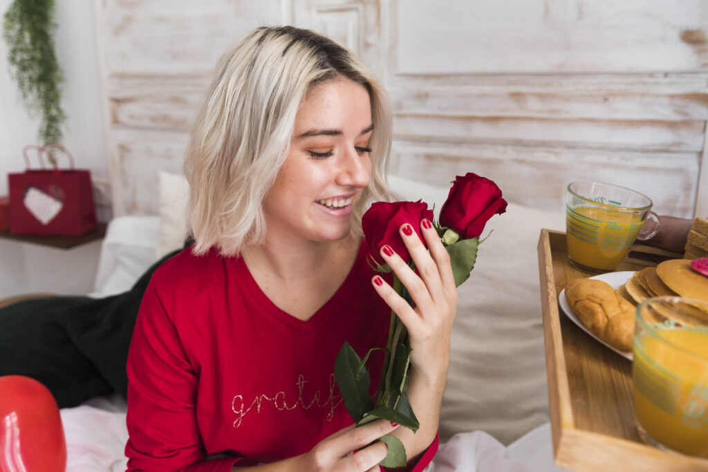 A woman holding red roses, sitting in bed with a breakfast tray.