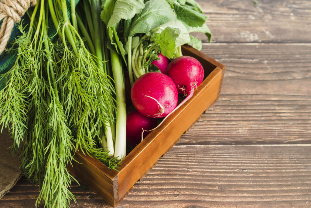 Fresh vegetables, including scallions, dill, and radishes, arranged in a wooden tray on a rustic surface.