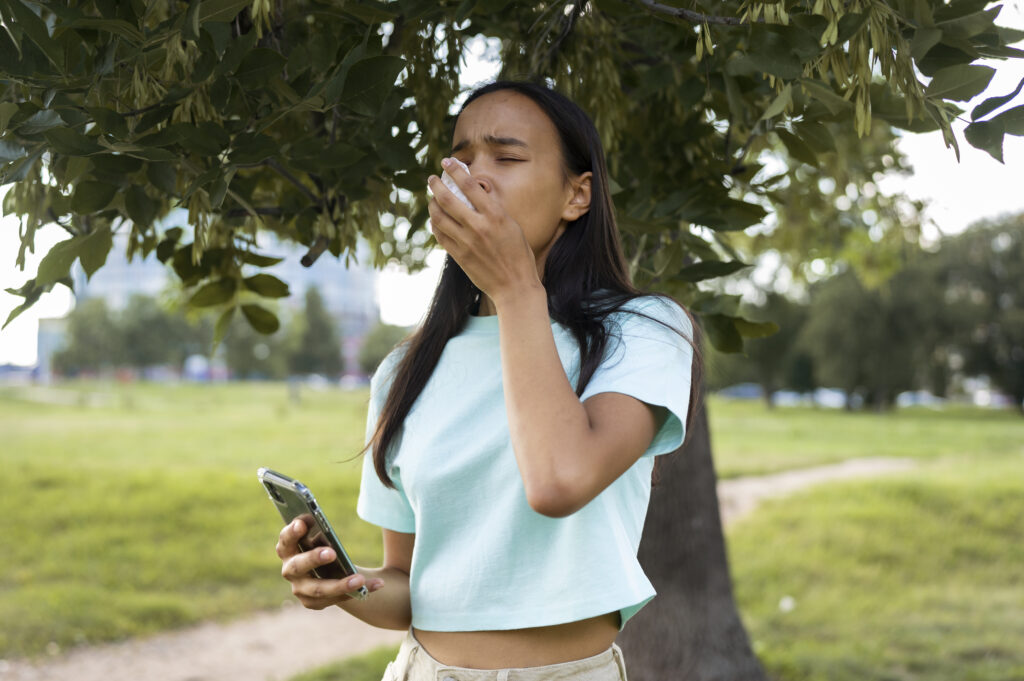 A woman outdoors holding her nose, showing discomfort from allergies.