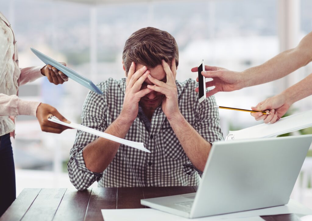 A stressed man sitting at a desk with his head in his hands while multiple hands offer him documents, a phone, and a pencil.