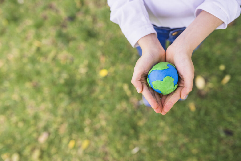 A child’s hands holding a small clay globe with green and blue representing Earth, with grass in the background.