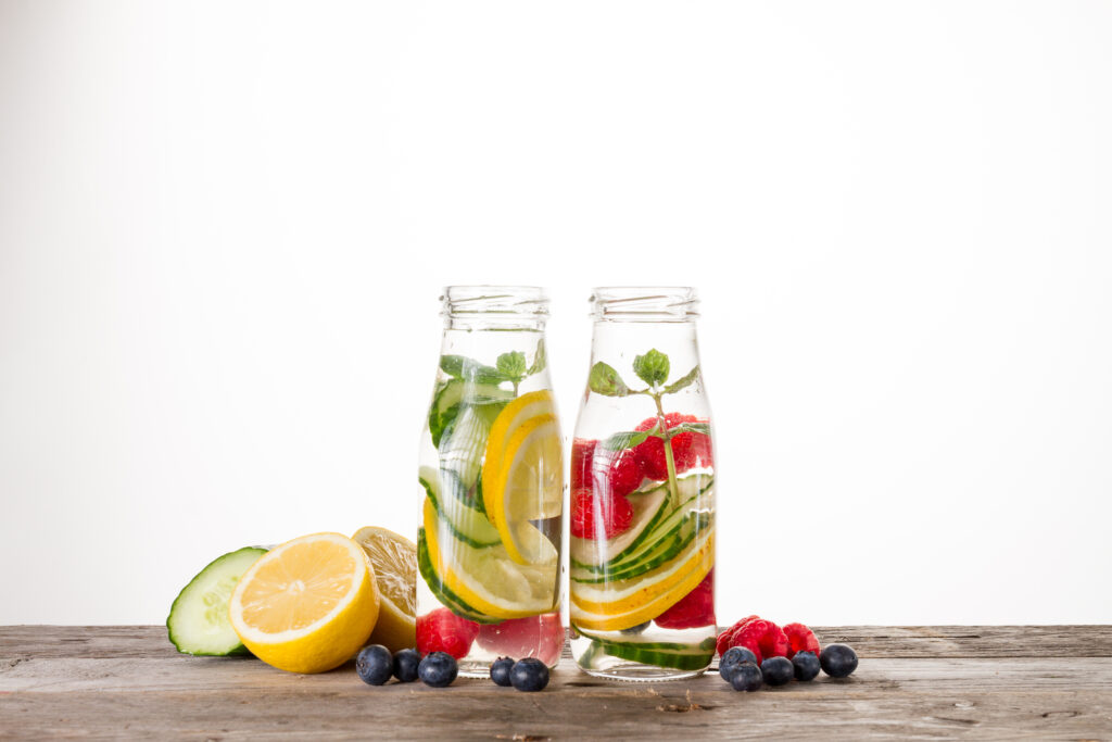 Two glass bottles of infused water with fruits like lemons, cucumbers, raspberries, and mint, placed on a wooden surface.